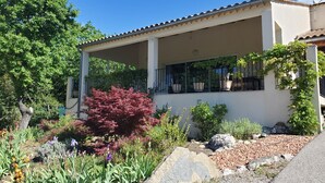 A two-story house with a covered patio, surrounded by a garden with various plants and a large rock. - Peaceful home with large covered south-facing terrace (Saint-Hippolyte-du-Fort)