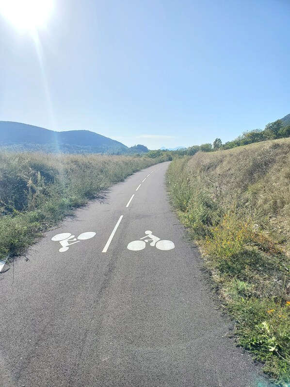 A paved path with bicycle symbols, flanked by grass and shrubs, leading towards distant hills. - Peaceful home with large covered south-facing terrace (Saint-Hippolyte-du-Fort)
