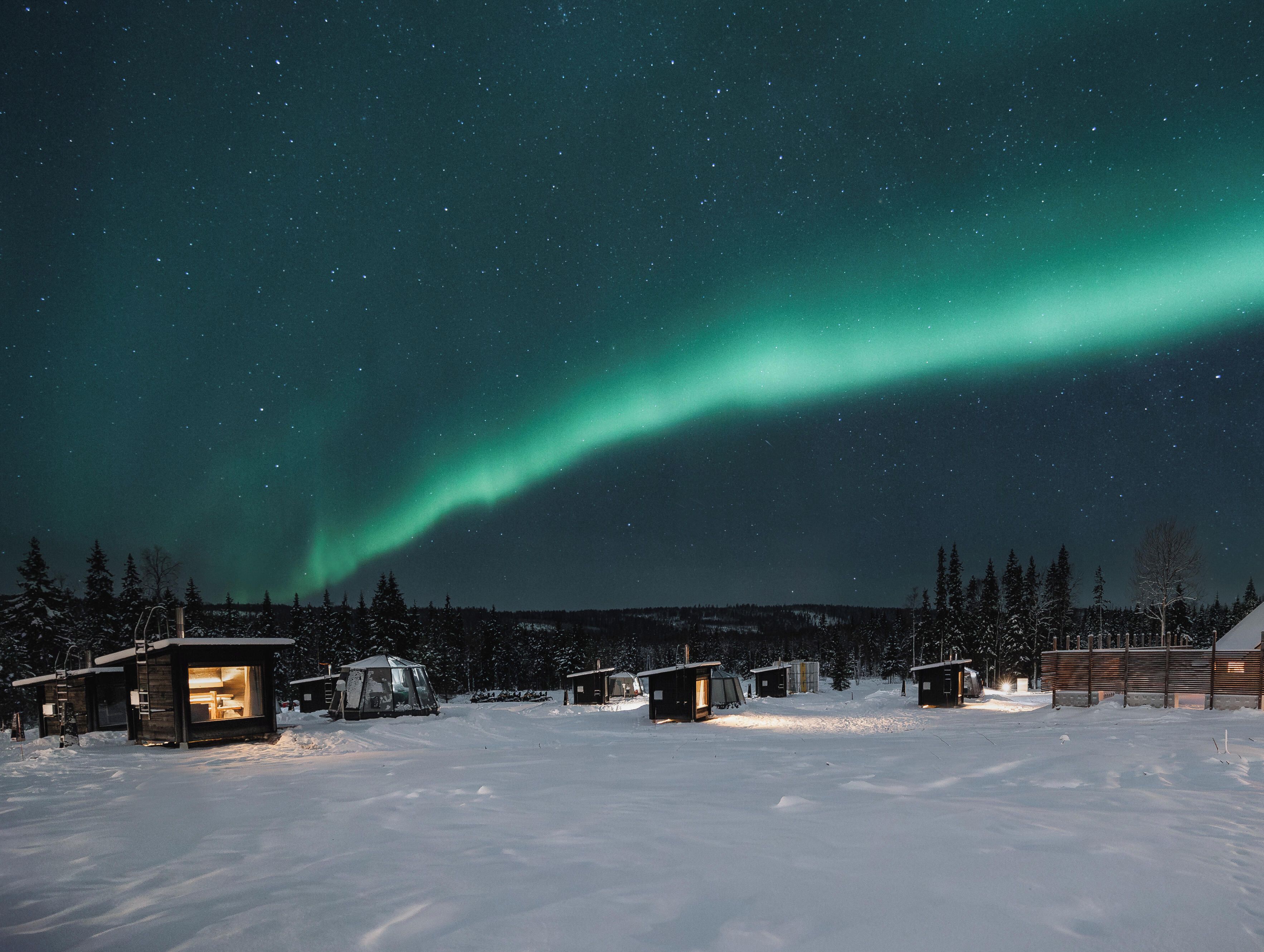 Paisaje nevado con una hilera de cabañas y una aurora boreal visible en el cielo nocturno.