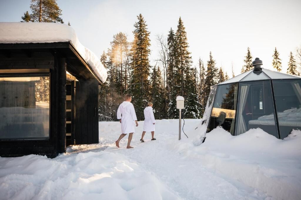 Una cabaña con cerramiento de vidrio y techo cubierto de nieve, rodeada de árboles.