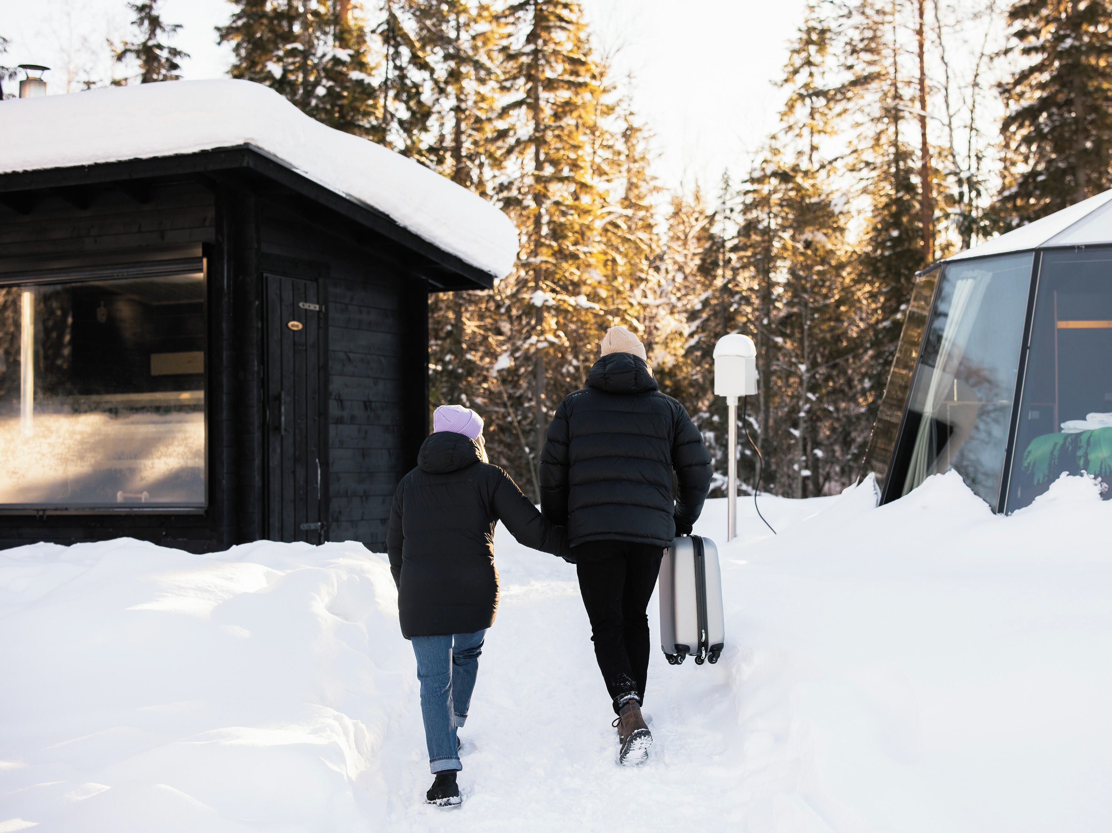 Dos personas caminando sobre un sendero cubierto de nieve hacia una cabaña con puerta de cristal, rodeada de árboles.