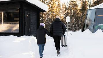 Dos personas caminando sobre un sendero cubierto de nieve hacia una cabaña con puerta de cristal, rodeada de árboles.