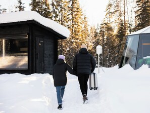 Zwei Personen gehen auf einem mit Schnee bedeckten Pfad zu einer Hütte mit Glastür, umgeben von Bäumen.