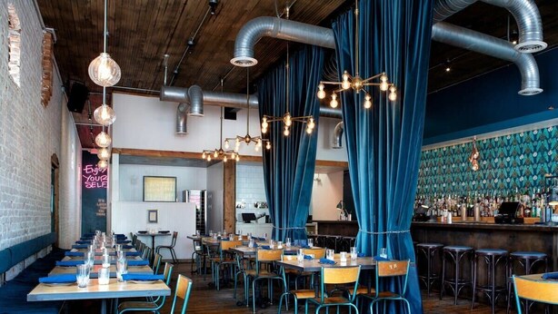 A restaurant with long communal tables, blue curtains, and a bar area with stools. - Splash Pad (Atlanta)