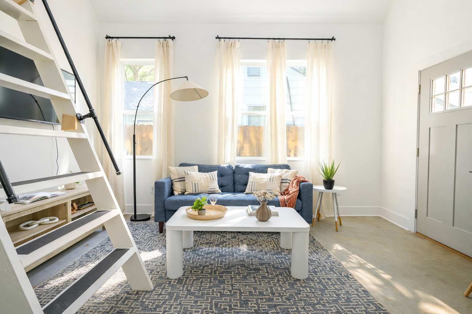 Una sala moderna con un sofá azul, una mesa de centro blanca y una alfombra estampada. Se ve una escalera a la izquierda, una lámpara de pie y una planta en maceta sobre una mesita.