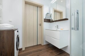 A modern bathroom with a wooden door, a washing machine, a sink with a mirror, and a glass shower enclosure.