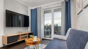 A modern living room with a grey sofa, a wooden TV stand with a flat-screen TV, a small round coffee table, and a sliding glass door leading to a balcony. - Stylish Apartment with Balcony | Parking in Garage (Świnoujście)