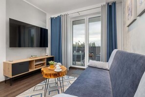 Ein modernes Wohnzimmer mit grauem Sofa, einem HolztV-Schrank mit Flachbildfernseher, einem kleinen runden Couchtisch und einer Glastür, die auf einen Balkon führt.