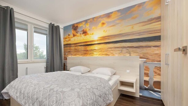 A bedroom with a bed, a nightstand, a window with curtains, and a mural of a sunset over the ocean. - Stylish Apartment with Balcony | Parking in Garage (Świnoujście)