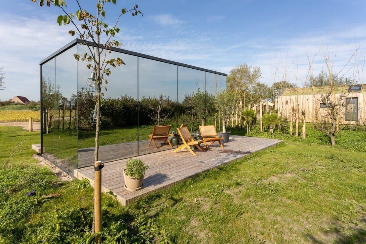 A modern glass structure with a wooden deck, outdoor seating, and a potted plant.