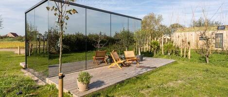 A modern glass structure with a wooden deck, outdoor seating, and a potted plant.