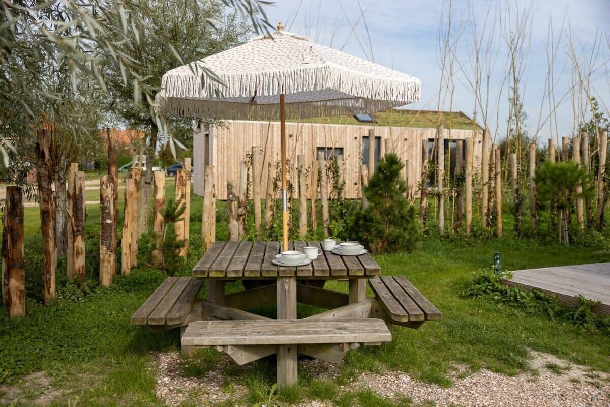 A wooden picnic table with a white umbrella, surrounded by a wooden fence and greenery.