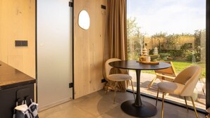 A modern room with a wooden floor, a round table with chairs, a view of a garden, and a sliding door.