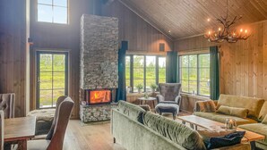 A cozy living room with a stone fireplace, wooden walls, and large windows offering a view of the outdoors. - Nice home in Trysil with house a mountain view (Trysil)