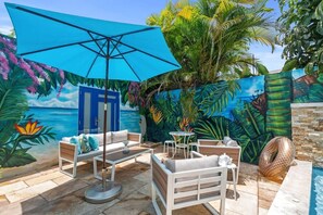 Un área de estar al aire libre con una sombrilla azul, muebles blancos y un mural de escena tropical.