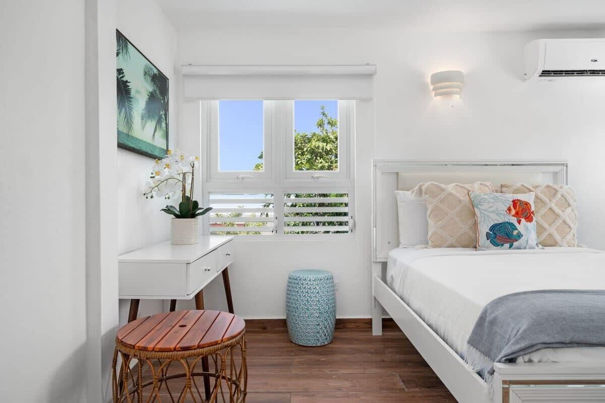 A bedroom with a bed, a desk, a chair, a window with blinds, and a small stool.