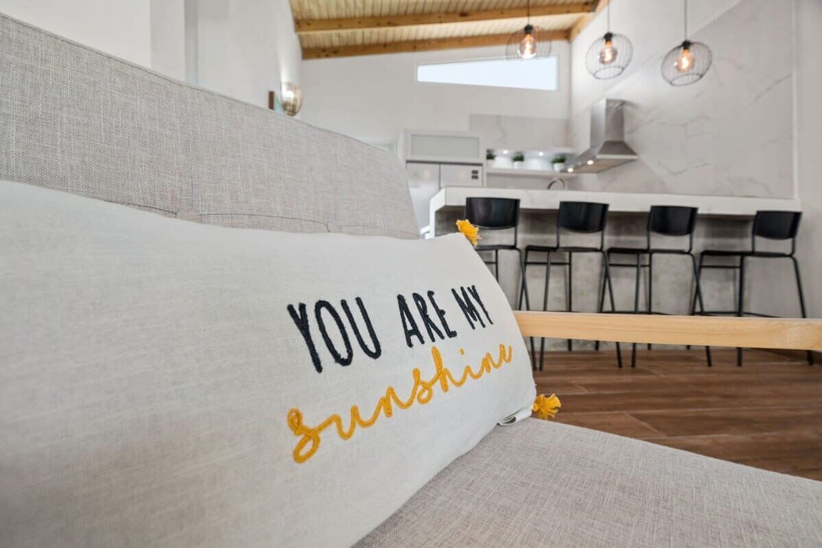 A modern living room with a sofa, a kitchen in the background, and a bar area with chairs.