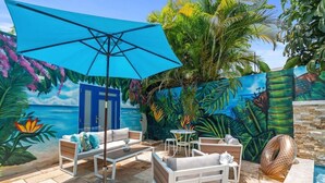 Un espace extérieur aménagé avec des sièges, un parasol bleu, du mobilier blanc et une fresque représentant un paysage tropical.