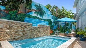 Une piscine ornée d’une fresque représentant un paysage tropical, un mur en pierre et un parasol bleu.
