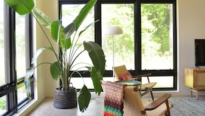 Quarto com janelas amplas, um vaso de planta, uma cadeira de fibra natural, uma mesa pequena e um abajur.