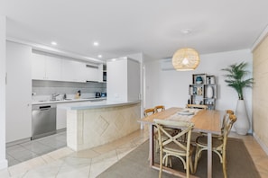 A modern kitchen with white cabinets, a built-in oven, and a dining area with a wooden table and chairs. - Apartment M204 - coastal and comfortable! (Scarborough)