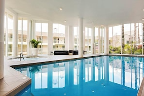 An indoor swimming pool with clear glass walls, a lounge area with seating, and a view of the surrounding buildings. - Apartment M204 - coastal and comfortable! (Scarborough)