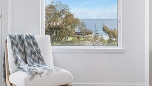 A room with a large window, a white chair with a blanket, and a view of the sea and trees. - Apartment M204 - coastal and comfortable! (Scarborough)