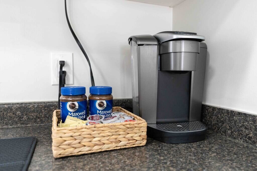 A coffee maker, two containers of Maxwell House coffee, and a basket on a kitchen counter.