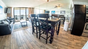 A spacious living area with a wooden floor, a dining table with chairs, a sofa, a ceiling fan, and a kitchen area with a refrigerator. - Garden City Beach Oceanfront Condo 807 | Rising Tides Properties Rental (Murrells)