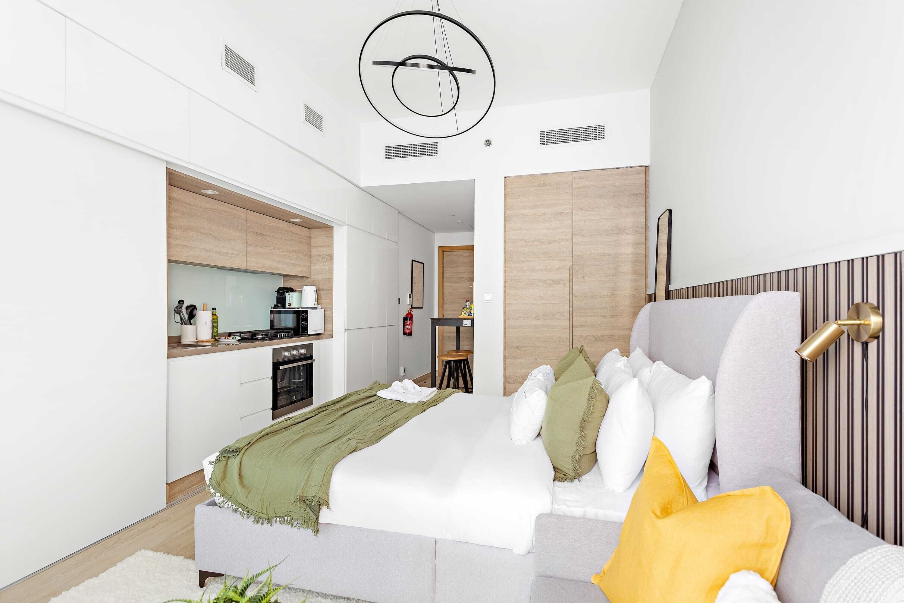 A modern hotel room with a bed, a sofa, a kitchen area, and a dining table.