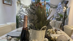 Una mesa de comedor adornada con una botella de champán, adornos plateados y dorados, y un pequeño árbol de Navidad.