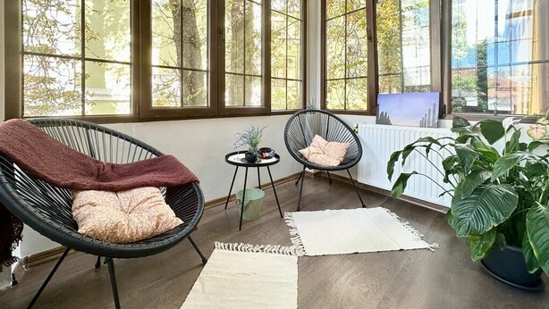 A room with large windows, a black chair, a small table with a plant, and a large potted plant. - Spacious 2-bed Flat  with Balcony, in central Bucharest - Universitate (București)