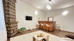 A modern hotel room with a wooden reception desk, three white bar stools, a flat-screen TV, a small round table with decorative items, a large window, and a patterned pillar. - Spacious 2-bed Flat  with Balcony, in central Bucharest - Universitate (București)