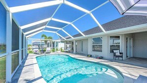 A covered swimming pool area with a clear sky, a pool, and a covered patio with chairs and a table.