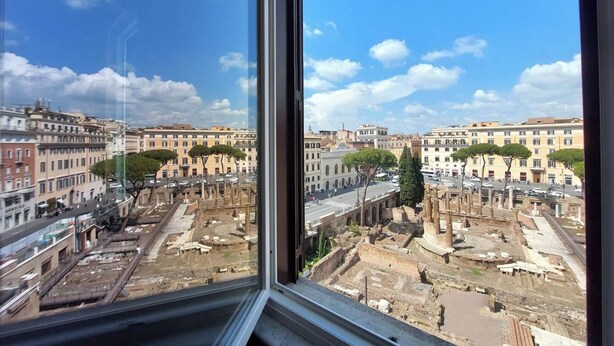 Une vue sur les ruines de la Rome antique, aperçues à travers une fenêtre, avec des bâtiments modernes en arrière-plan.