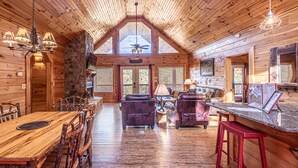 A spacious log cabin interior with a high ceiling, a dining area with a wooden table and chairs, a stone fireplace, and a bar area with stools.