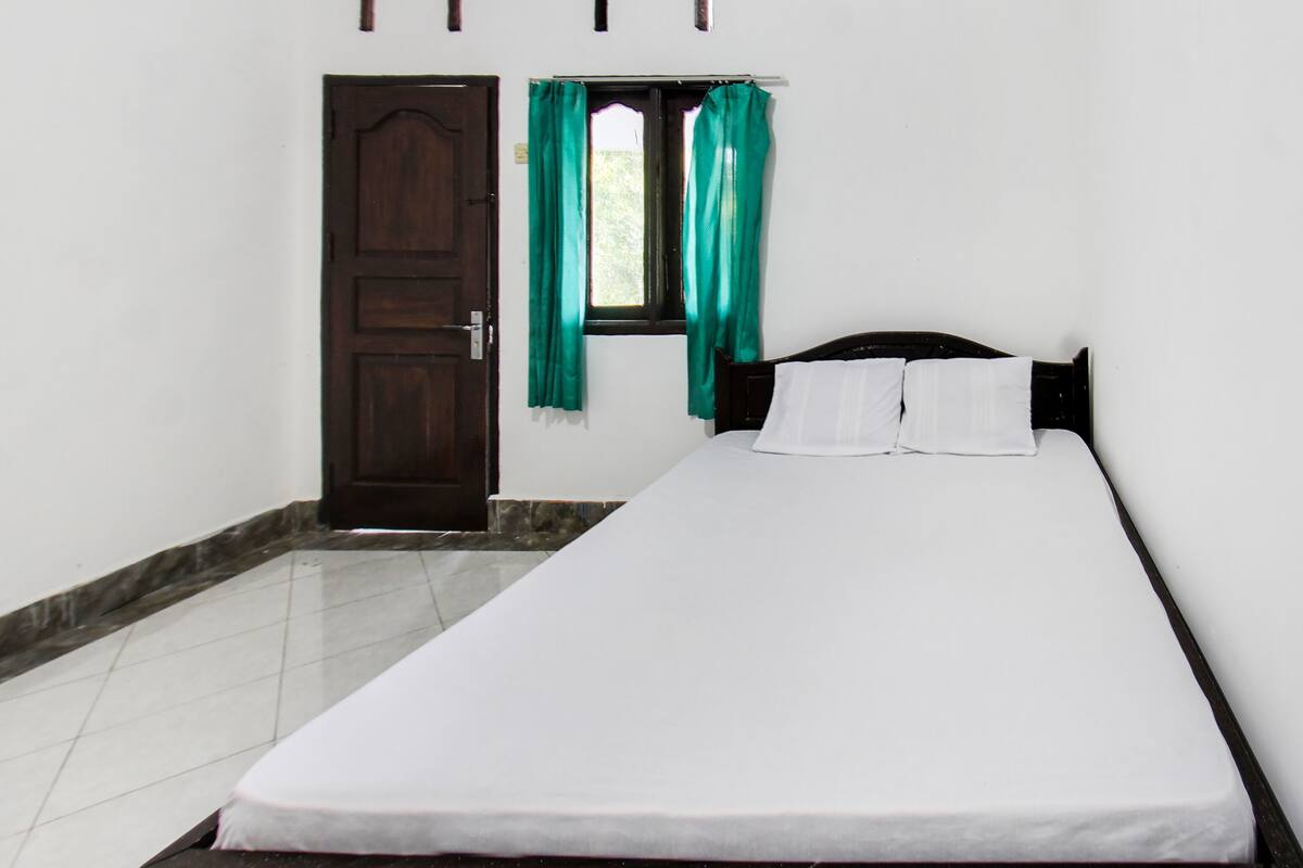 Een kamer met een enkel bed, een houten deur, een raam met groene gordijnen en een wit sprei.