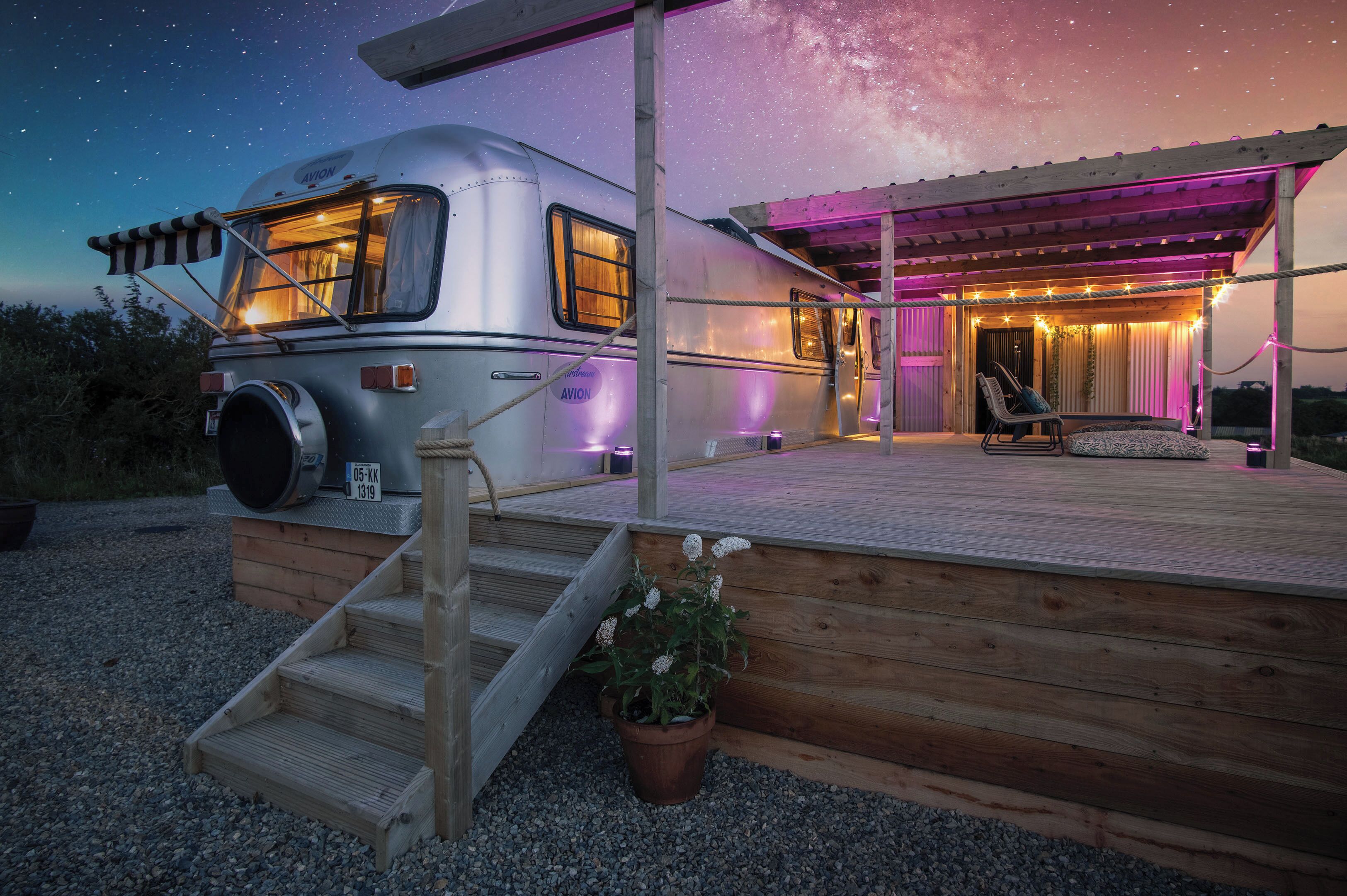 Ein stehender Wohnmobil mit einer Holzterrasse, einem überdachten Sitzbereich und einem sternenübersäten Nachthimmel.