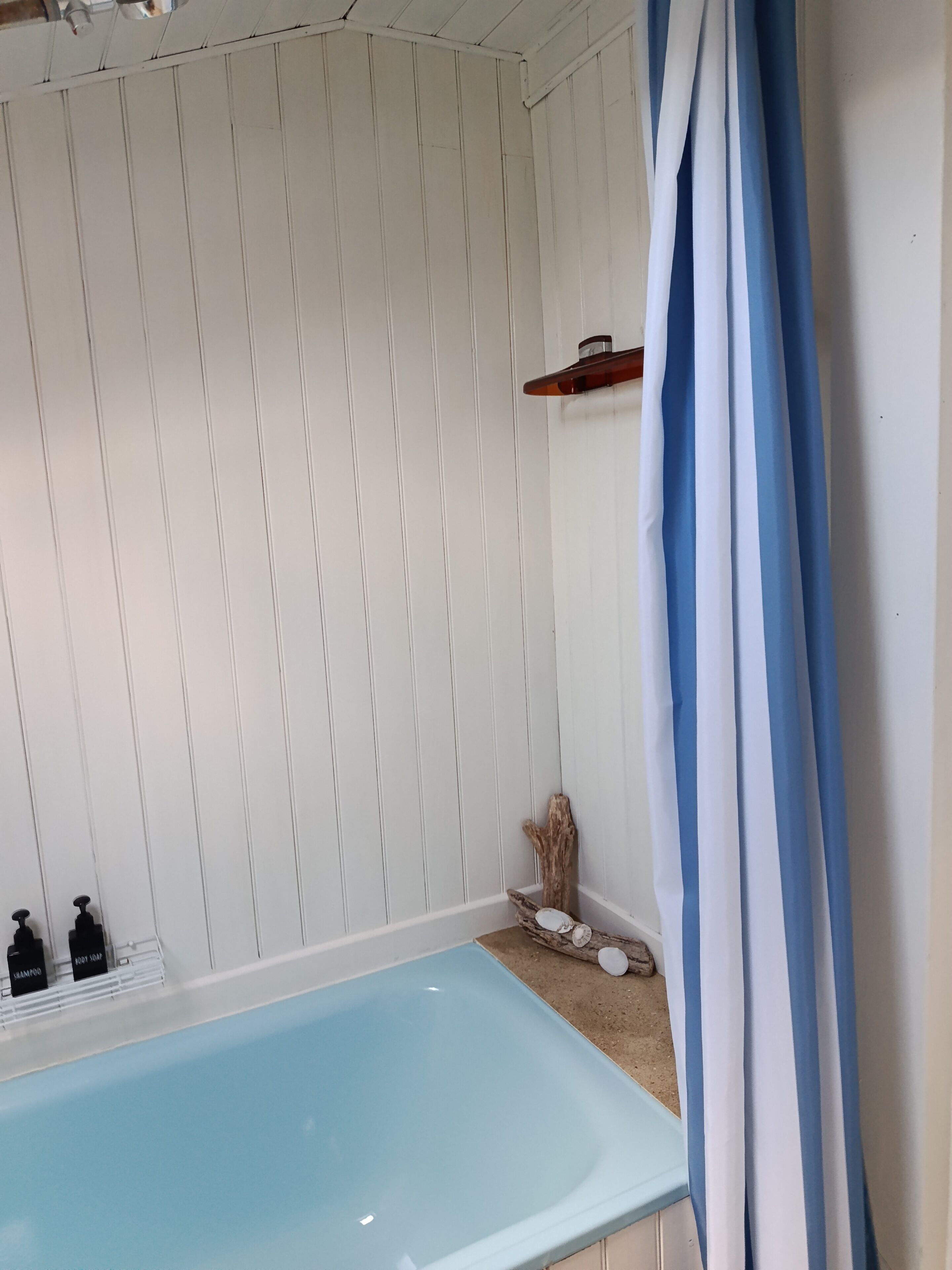 A bathroom with a bathtub, white wooden walls, a blue and white striped shower curtain, and a shelf with toiletries.
