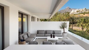 A modern balcony with a grey sectional sofa, white coffee table, and wicker chairs. A potted tree and two candle holders are on the table. The balcony overlooks a mountainous landscape with buildings in the background. - Exceptional 3 bedroom duplex penthouse in Epic Marbella (Marbella)