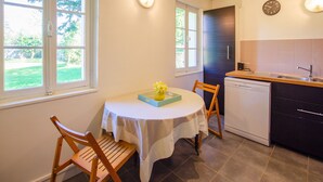 A kitchen with a dishwasher, a dining table with chairs, and two windows with a view of greenery. - L'If, ground floor flat in a beautiful house, close to the beach (Varengeville-sur-Mer)