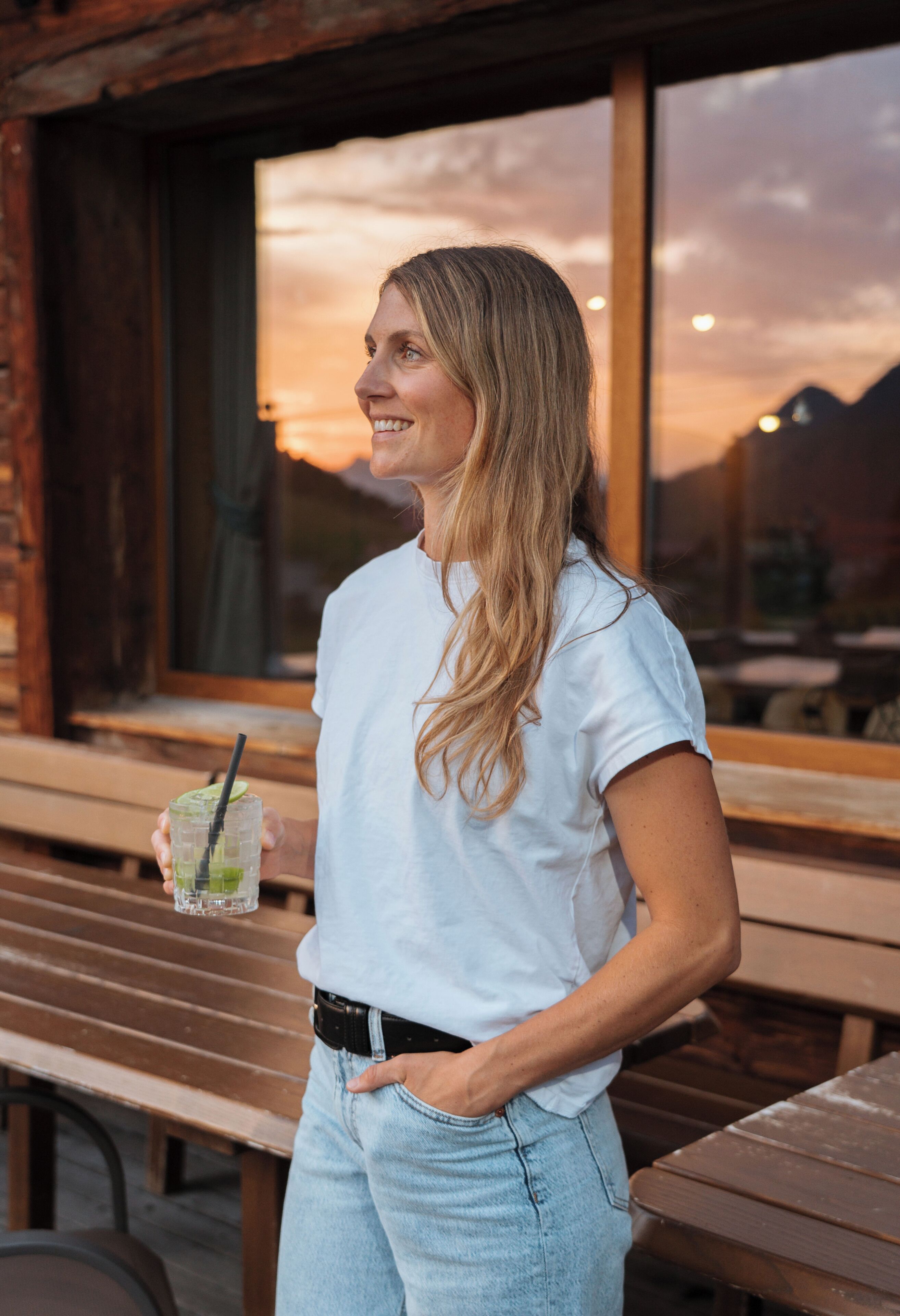 A person standing at a wooden table with a drink, overlooking a scenic view through a window.