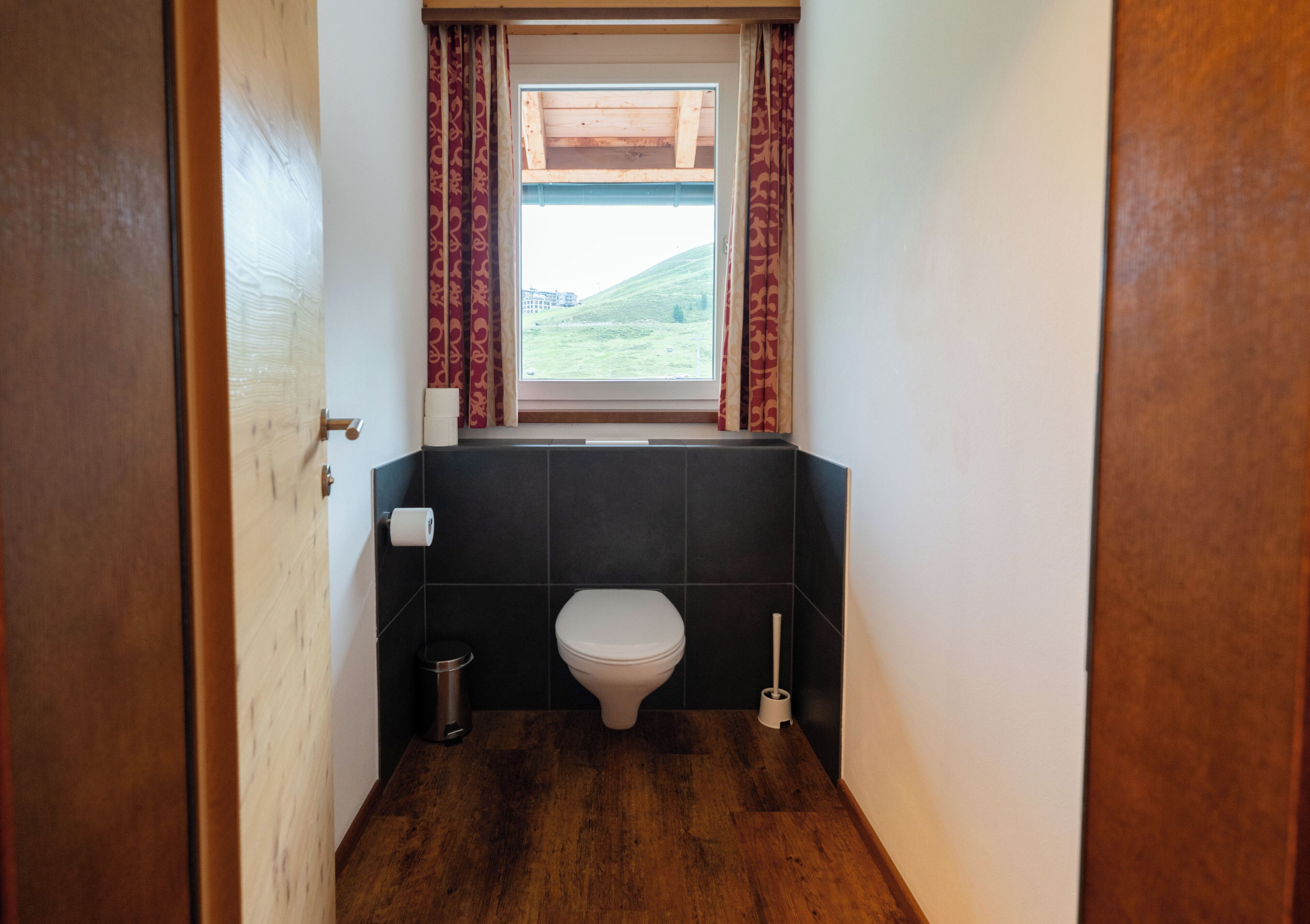 A narrow bathroom with a toilet, a bidet, and a window with curtains.