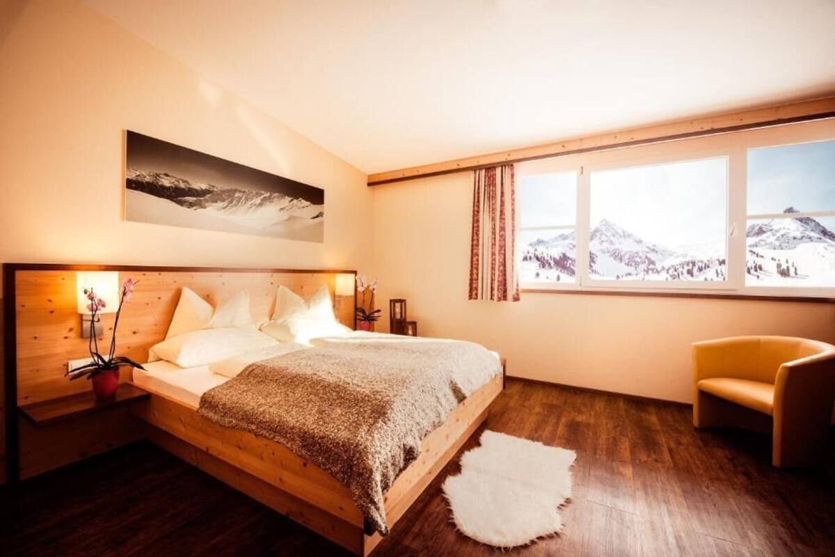 Un dormitorio con una cama de madera, vistas a montañas a través de ventanas amplias, una silla y una alfombra pequeña.