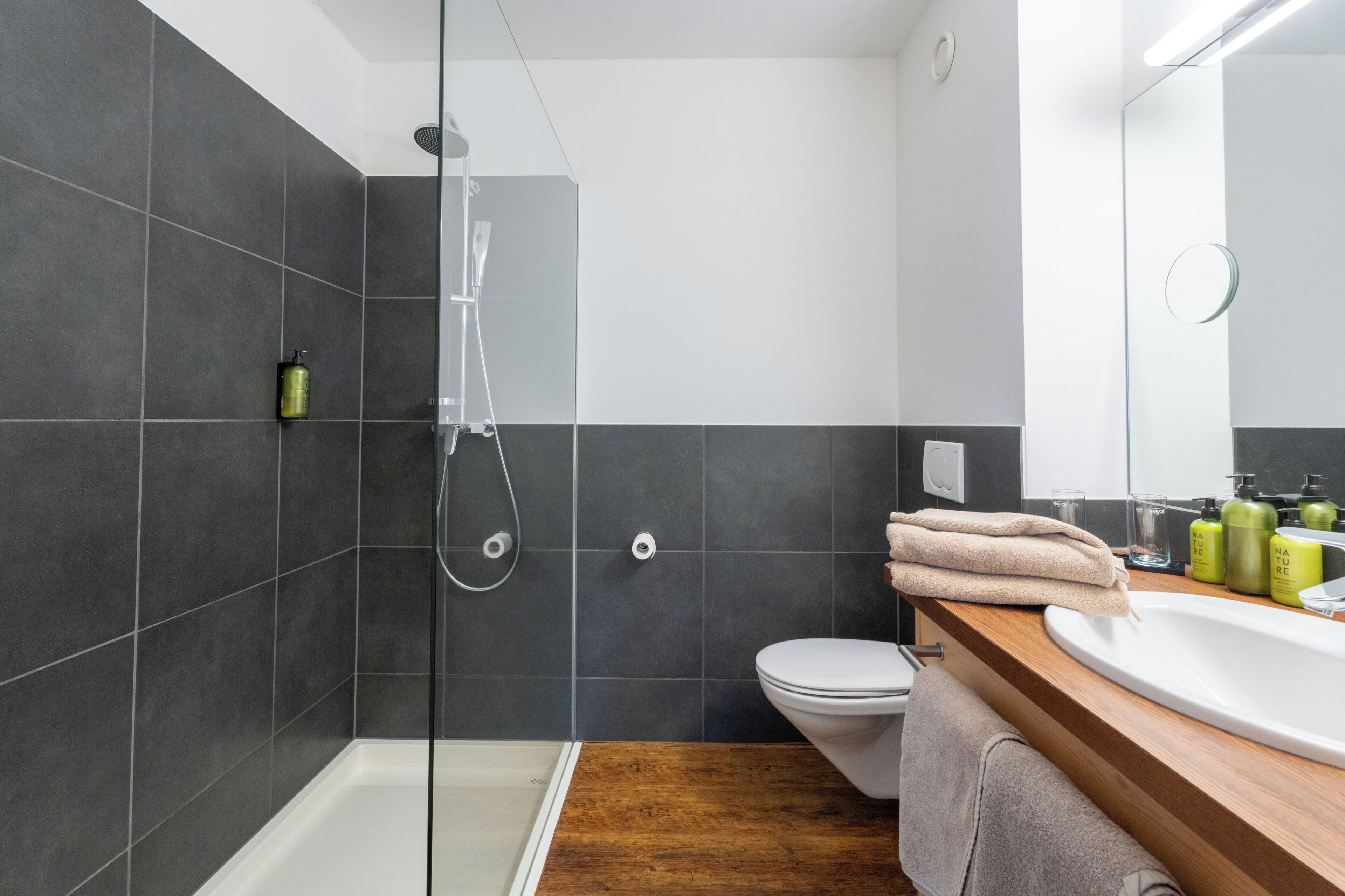 Un baño moderno con ducha cubierta por vidrio, inodoro blanco y tocador de madera con lavabo y espejo.