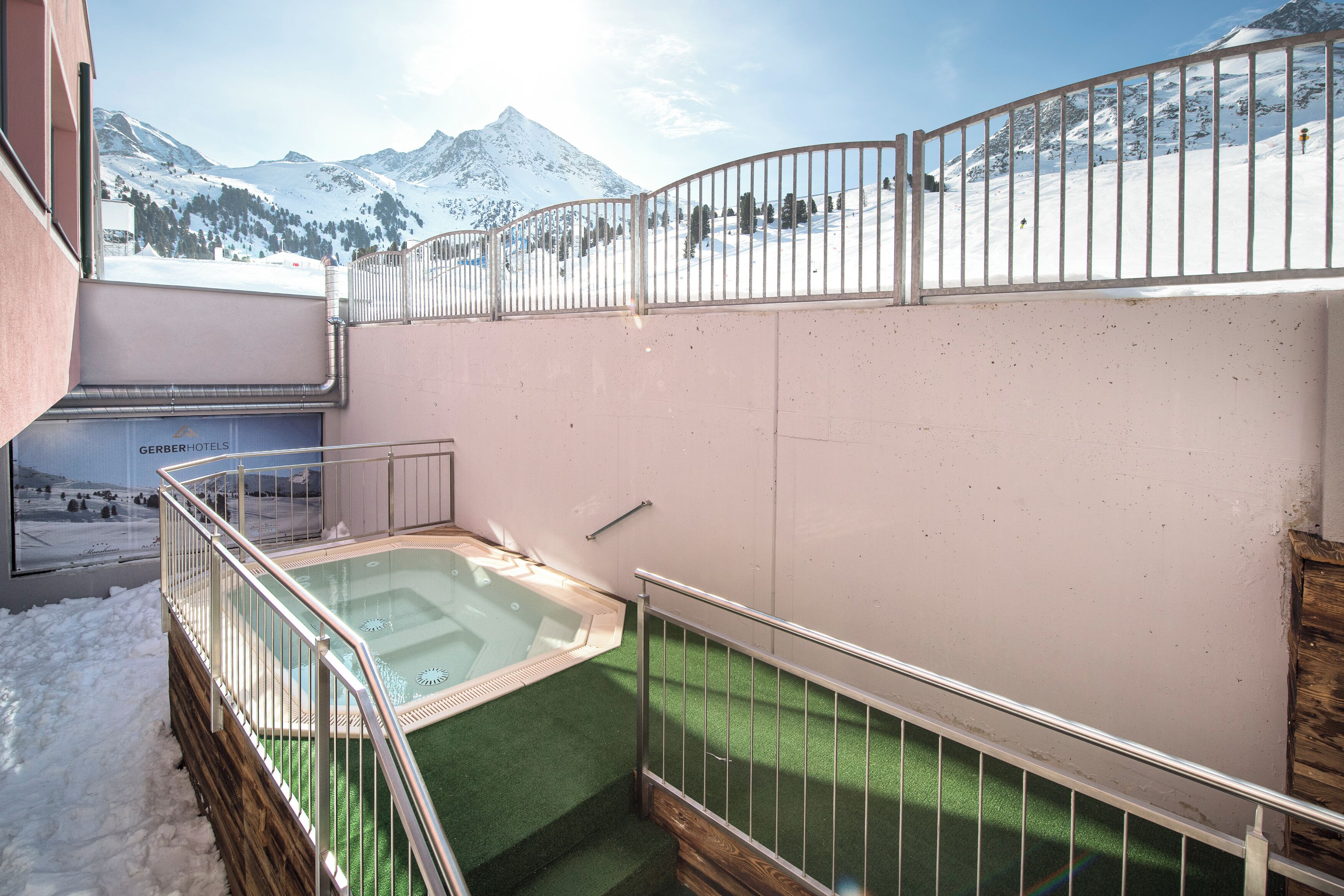Un área de spa con un jacuzzi, rodeado por una barandilla de metal, y vistas a montañas nevadas al fondo.