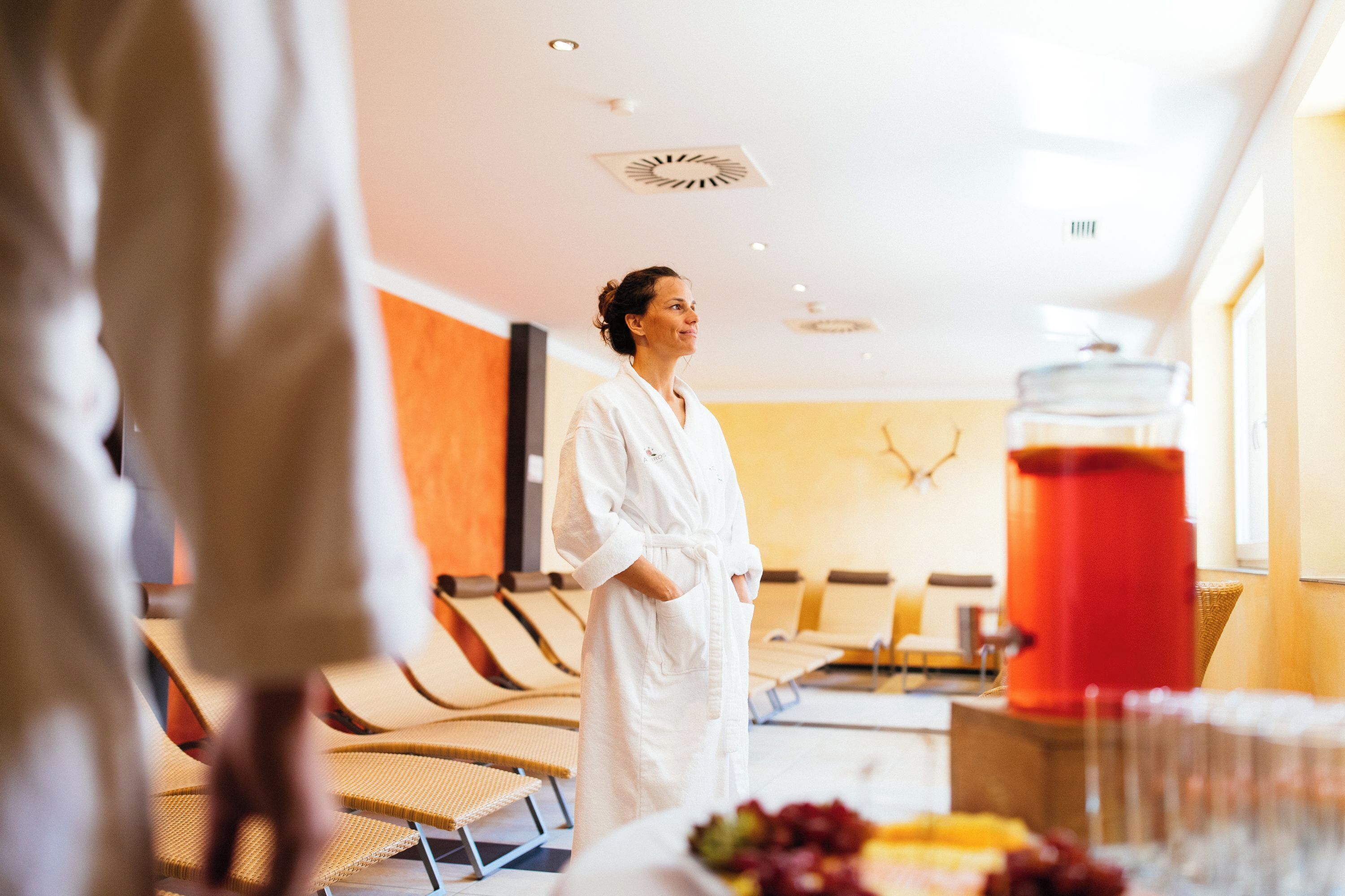 A person in a white robe standing in a room with a large red container, beige chairs, and a window.