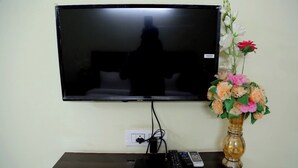Un televisor de pantalla plana montado en una pared, una regleta de enchufes, un control remoto y un jarrón con flores sobre una estantería.