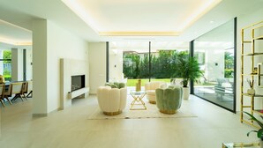 A modern living room with a flat-screen TV, two sofas, a coffee table, and a large window offering a view of a garden. - Magnificient 5 bed villa in Golden Mile Marbella (Marbella)