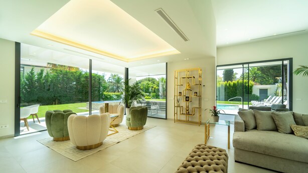 A modern living room with a large glass door leading to a pool area, a beige sofa, and a tufted ottoman. - Magnificient 5 bed villa in Golden Mile Marbella (Marbella)